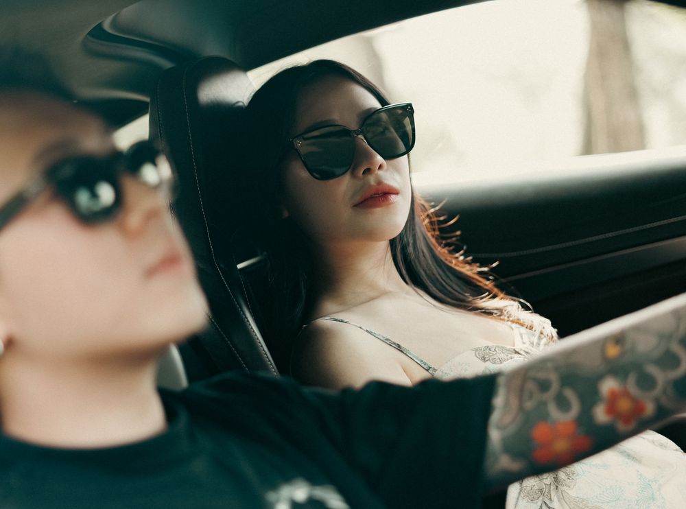 Two women wearing black sunglasses sitting inside a car, one with tattoos driving and one with dark hair in the passenger seat.