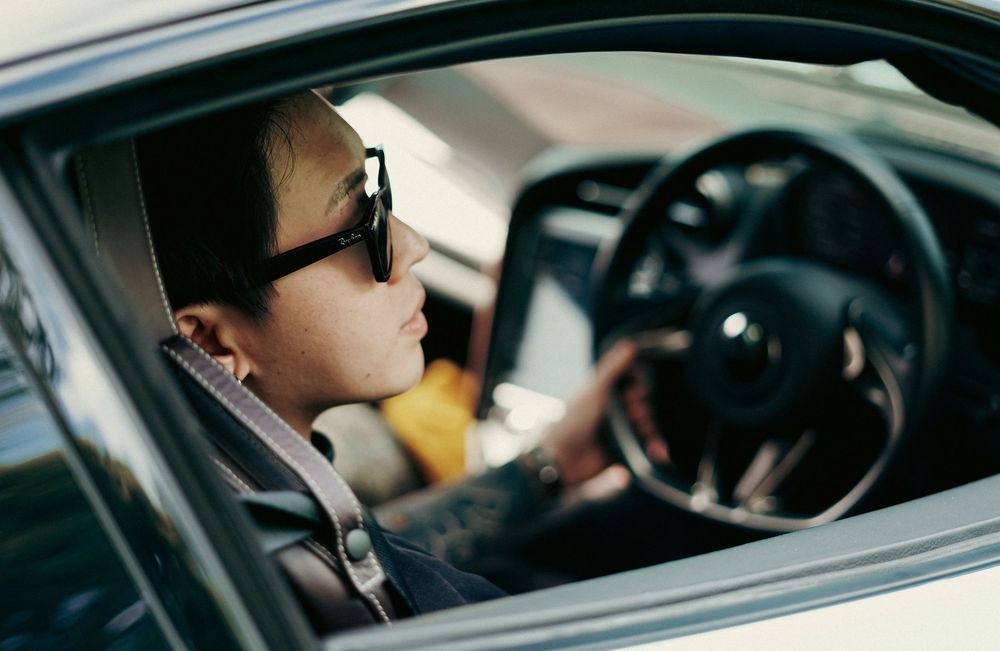 Tattooed person with sunglasses driving a luxury car, viewed through open window.
