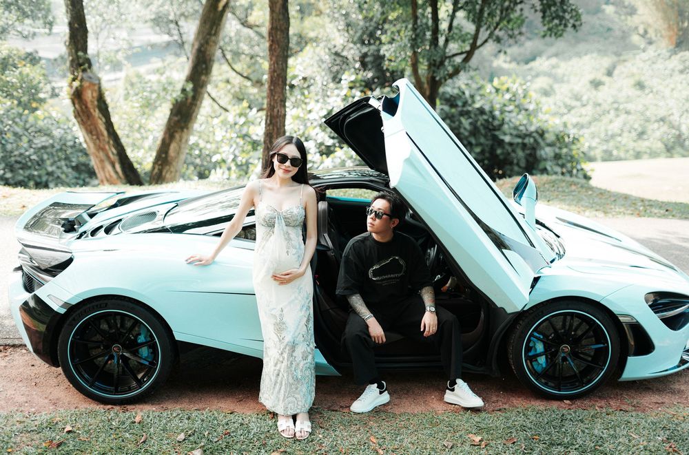 Pregnant woman in white lace dress poses with man beside teal McLaren supercar with scissor doors open outdoors.