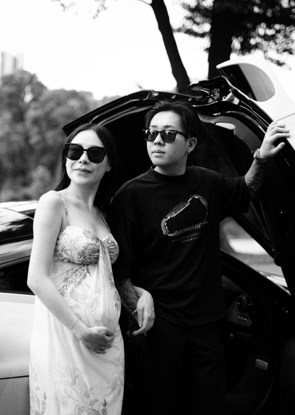 Black and white photo of a stylish couple wearing sunglasses posing near an open car trunk outdoors.