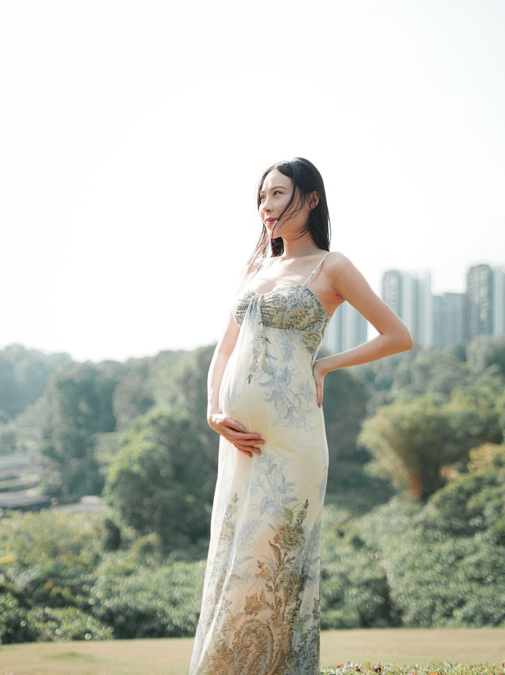 Pregnant woman in elegant floral maxi dress standing outdoors with lush greenery and city skyline in background.