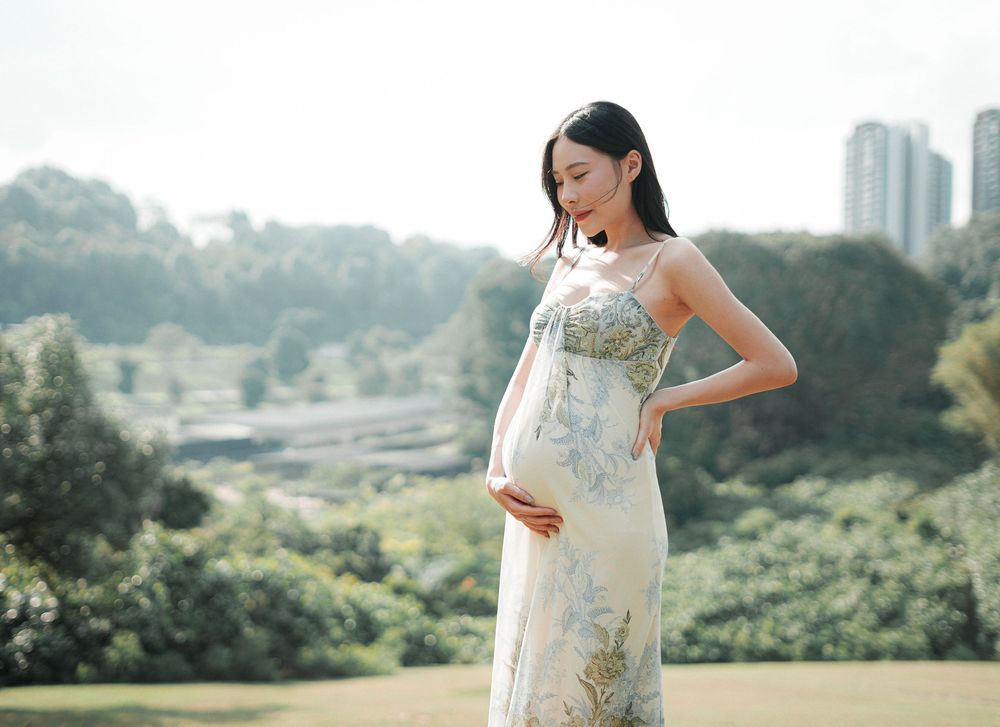 Pregnant woman in floral sundress cradling baby bump outdoors with lush greenery and city skyline in background.