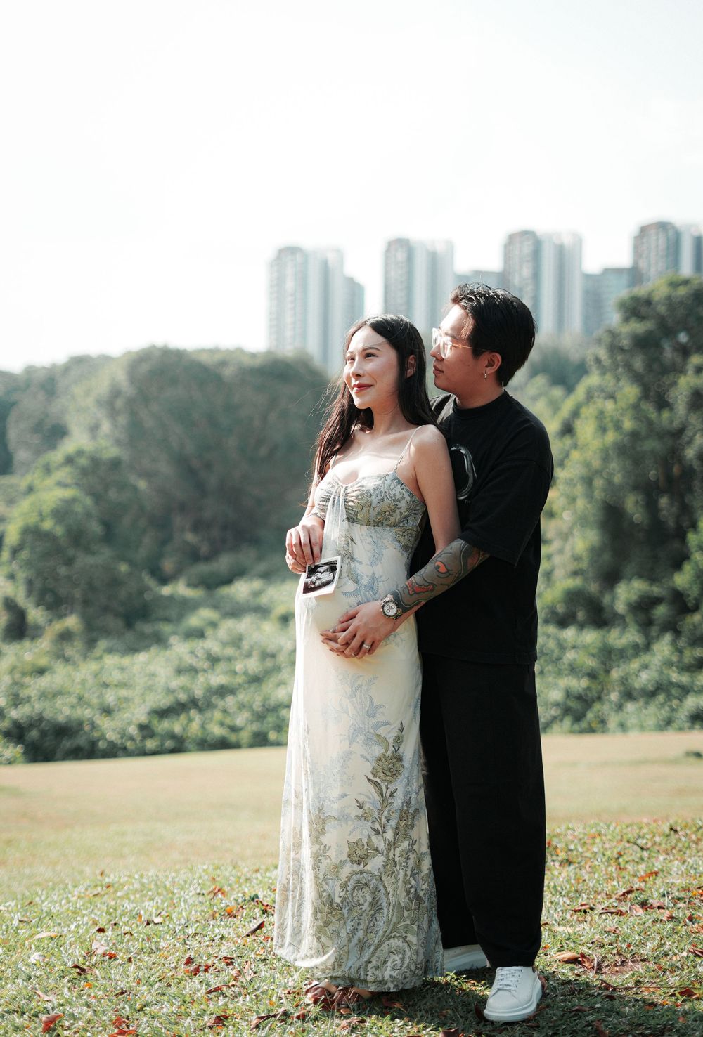 Couple poses outdoors on grass, woman in floral gown holding ultrasound, man in black embracing her, city skyline behind.