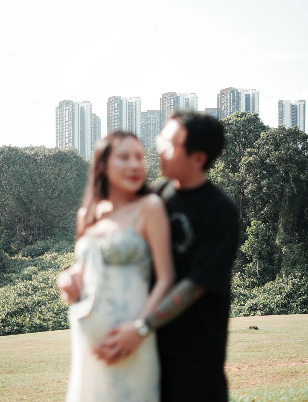 Couple posing together in soft focus with green trees and tall buildings behind them outdoors.