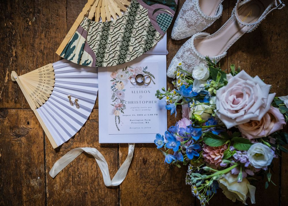 Wedding flat lay featuring floral bouquet, invitation, fans, earrings, and bridal heels on wood.