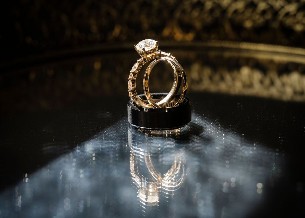 Elegant gold diamond engagement ring and wedding band set reflected on glossy dark surface.