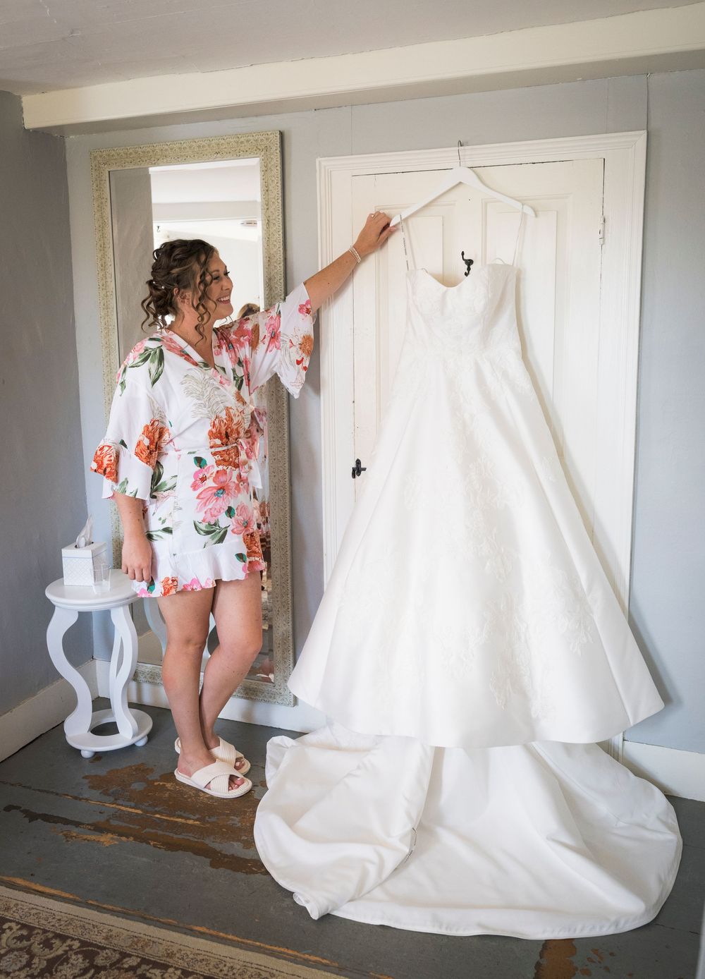 Bride in floral robe admiring her white wedding gown hanging on a door before her special day.