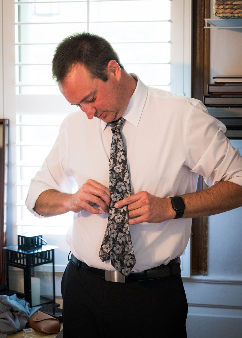 Groom buttoning his shirt while wearing a floral tie during wedding day preparations.