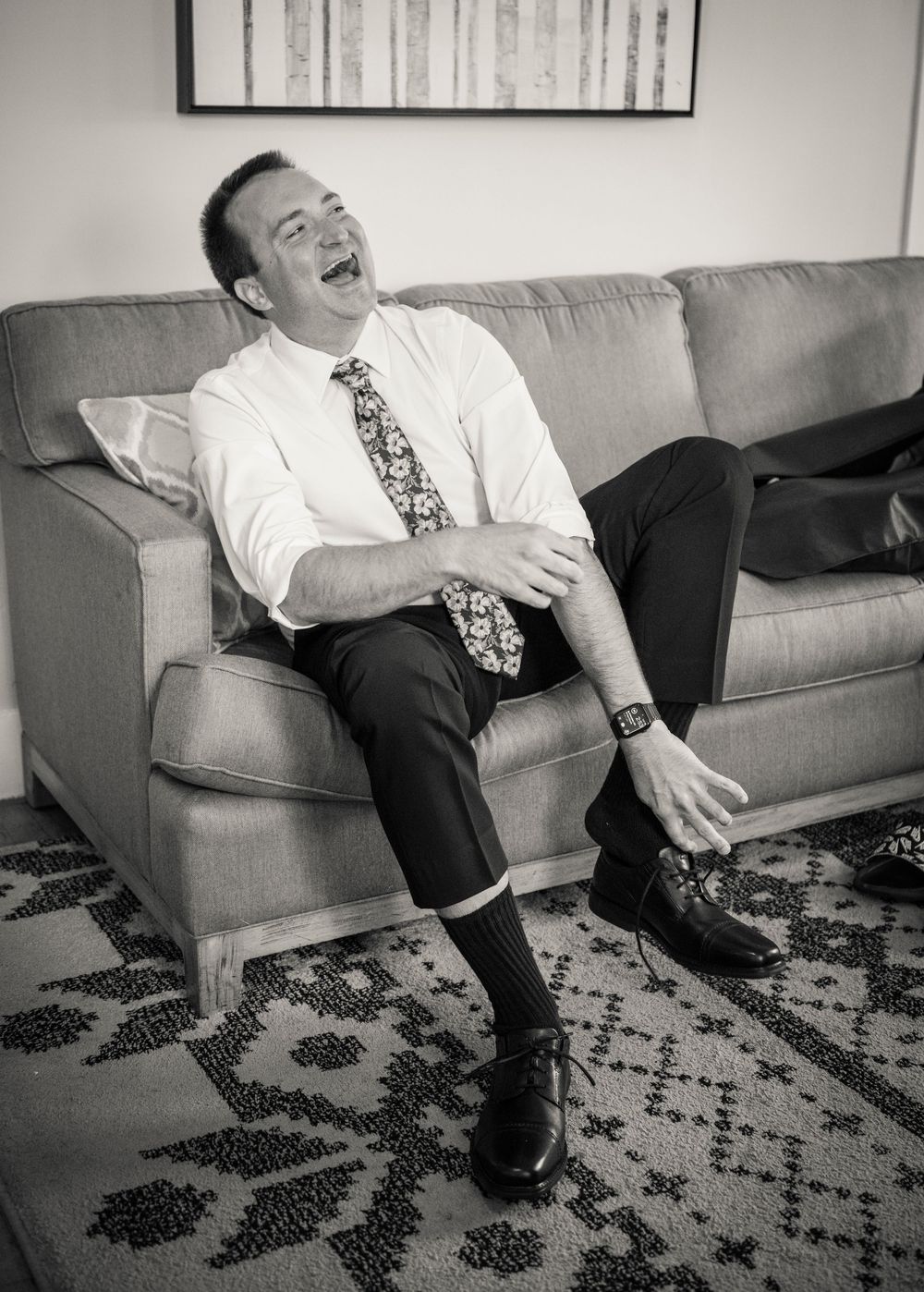 Groom laughing joyfully on a couch in black and white photo during wedding preparations.