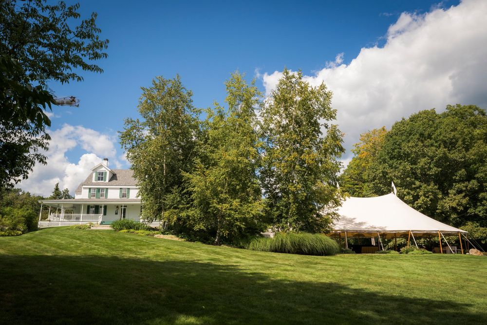 White wedding tent set up on lush green lawn beside a charming white farmhouse on sunny day.