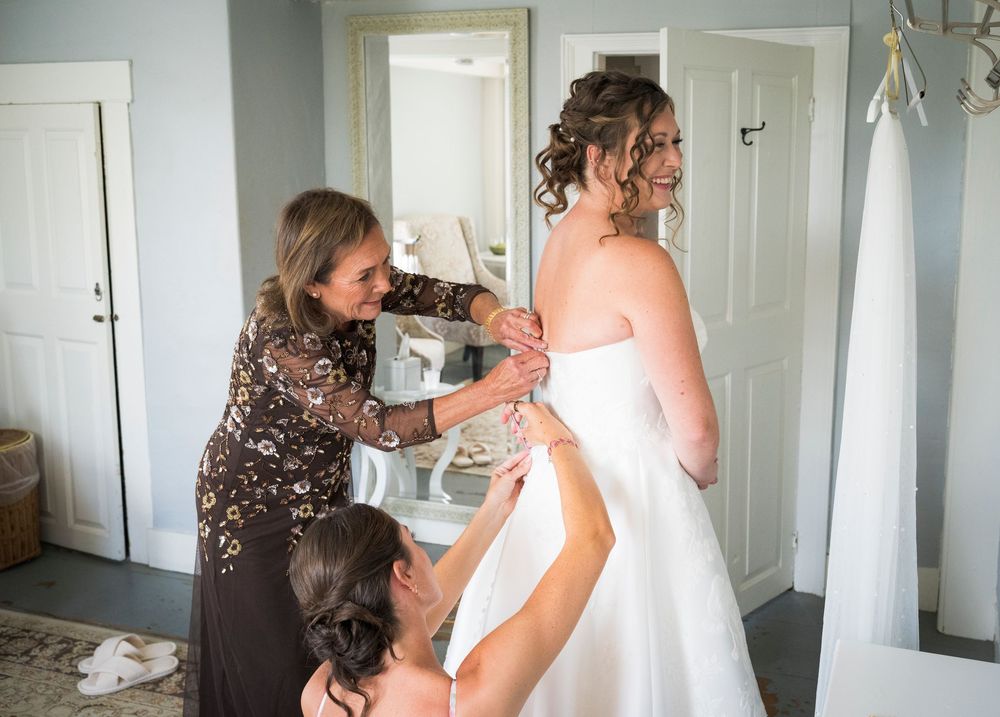 Bride smiles in mirror as mother and bridesmaid help fasten her strapless white wedding gown.