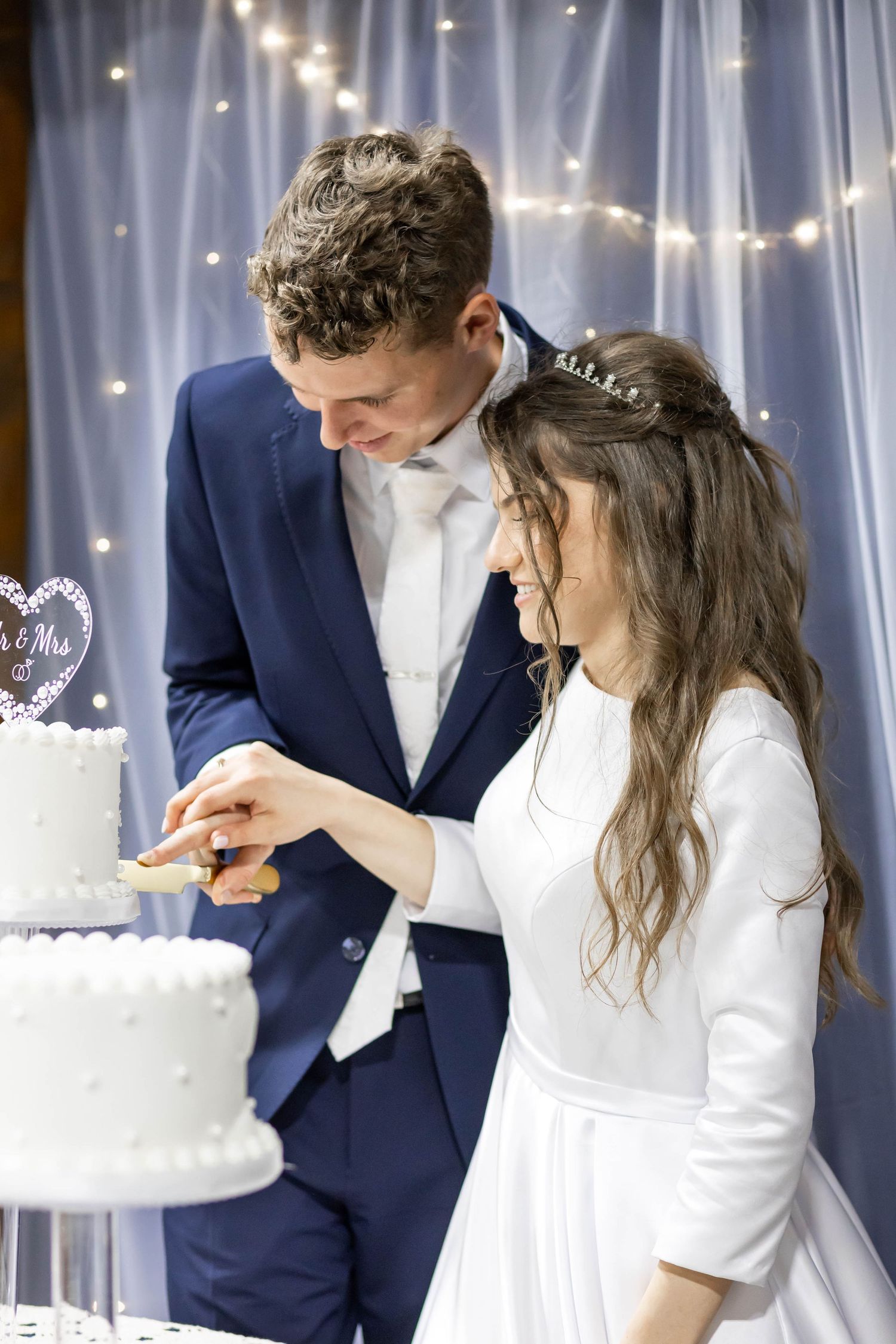Bride and groom cutting their white wedding cake together, smiling, with fairy lights in the background.