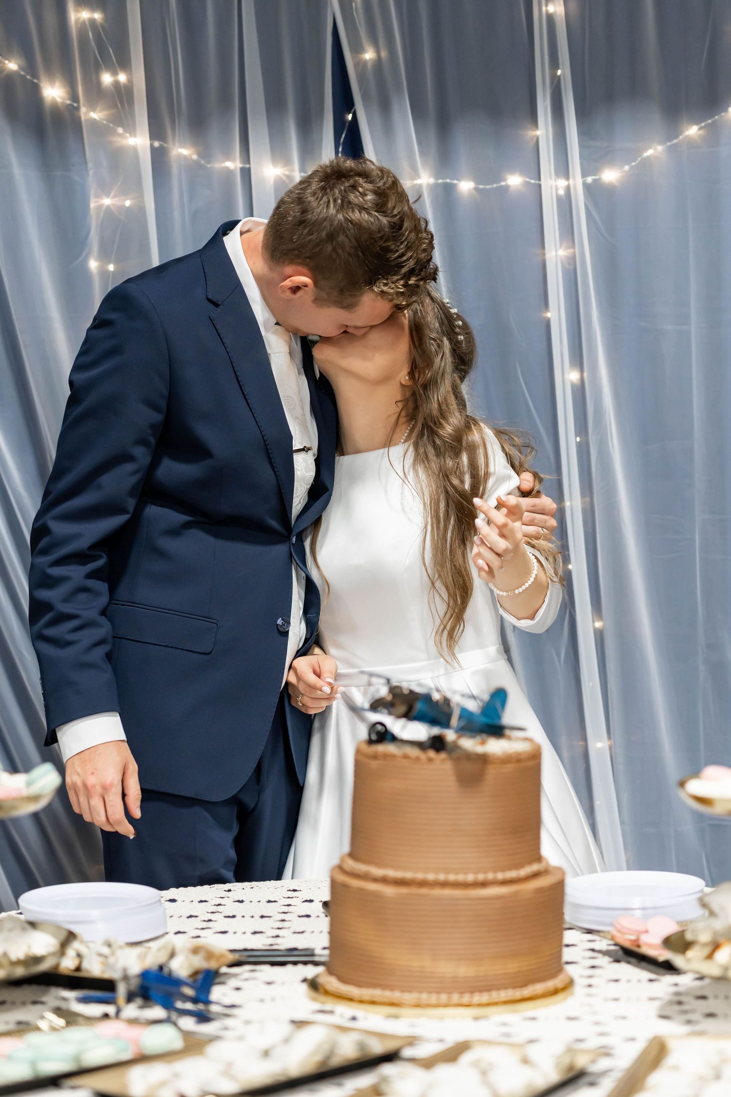 Bride and groom kissing while cutting a two-tier chocolate wedding cake decorated with a toy airplane topper.
