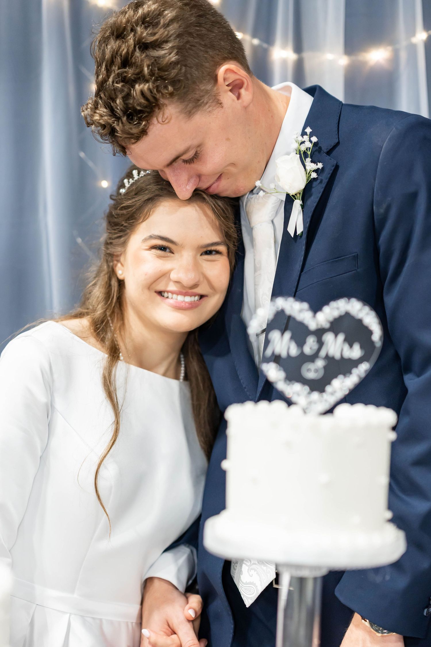 Smiling bride and groom in navy suit pose together near a white Mr. & Mrs. wedding cake with fairy lights backdrop.