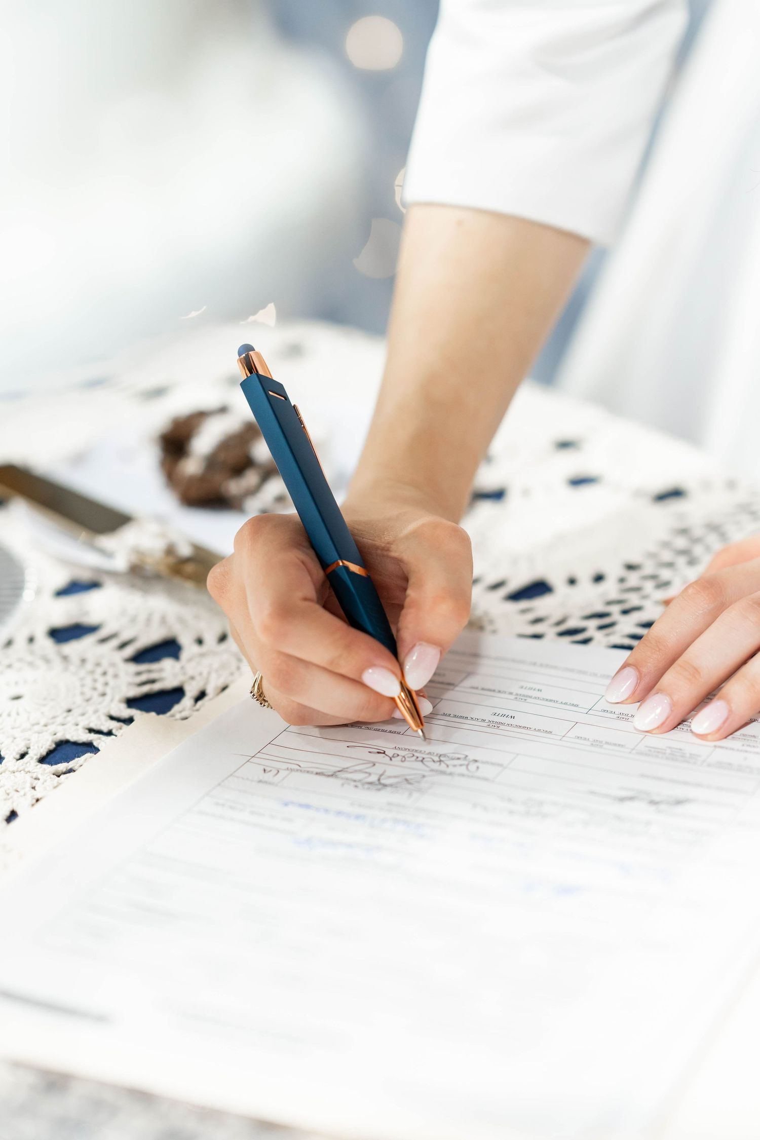 Close-up of a woman's hand writing on a document with a blue and gold pen on a lace tablecloth.