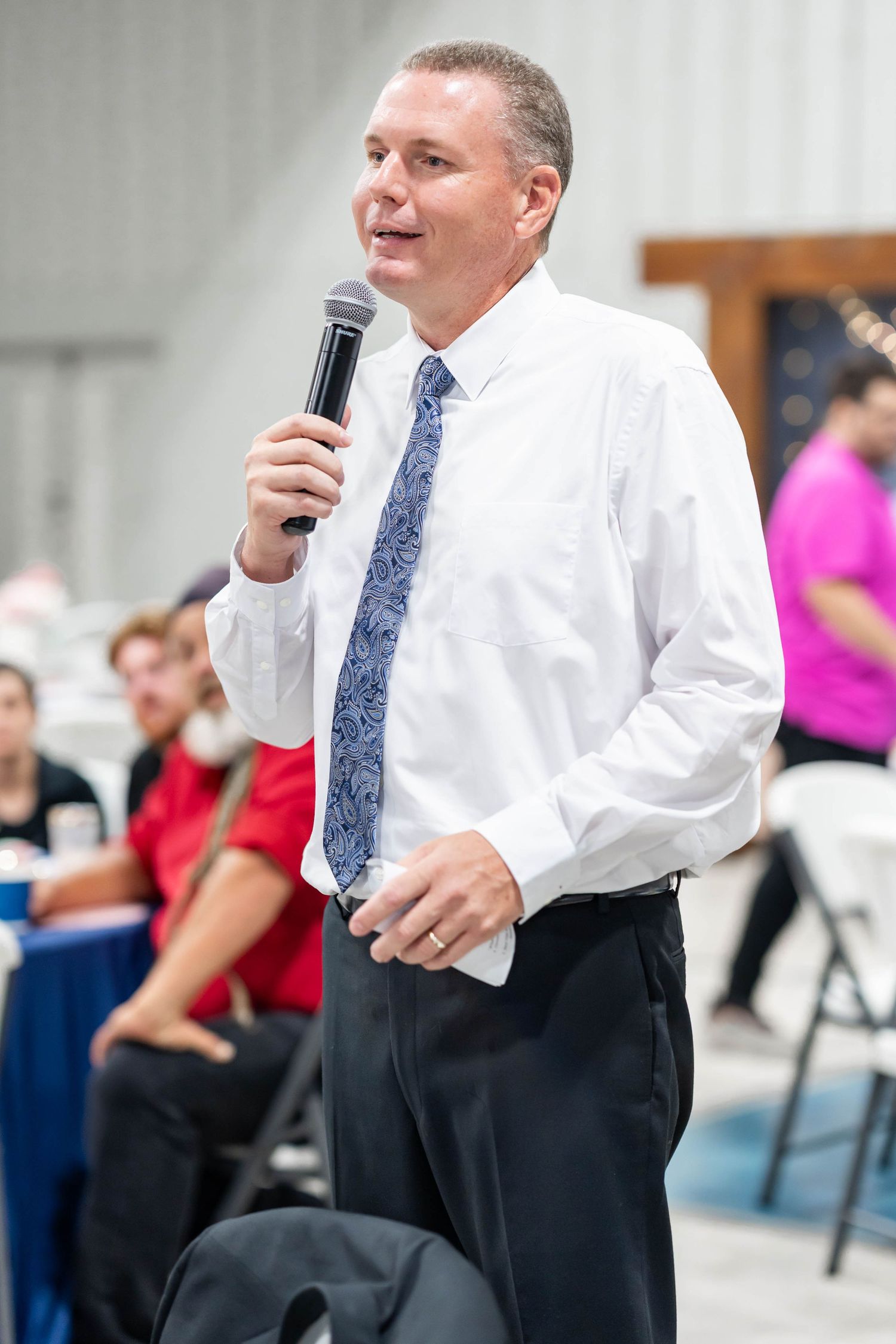 Man in white shirt and blue tie speaks into microphone at indoor event with seated audience in background.