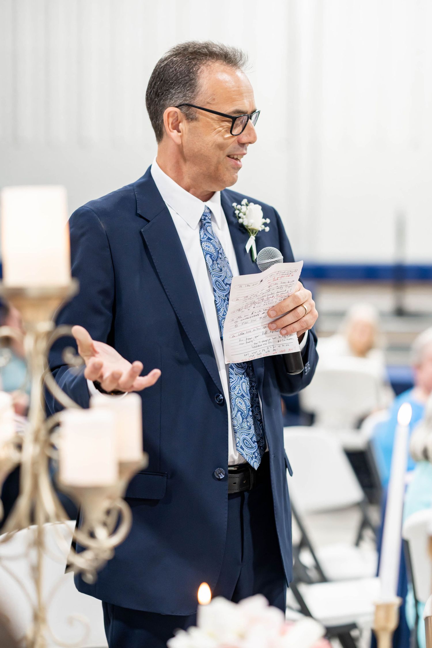 Man in navy suit with blue tie giving a speech at a wedding reception, holding notes and a microphone.