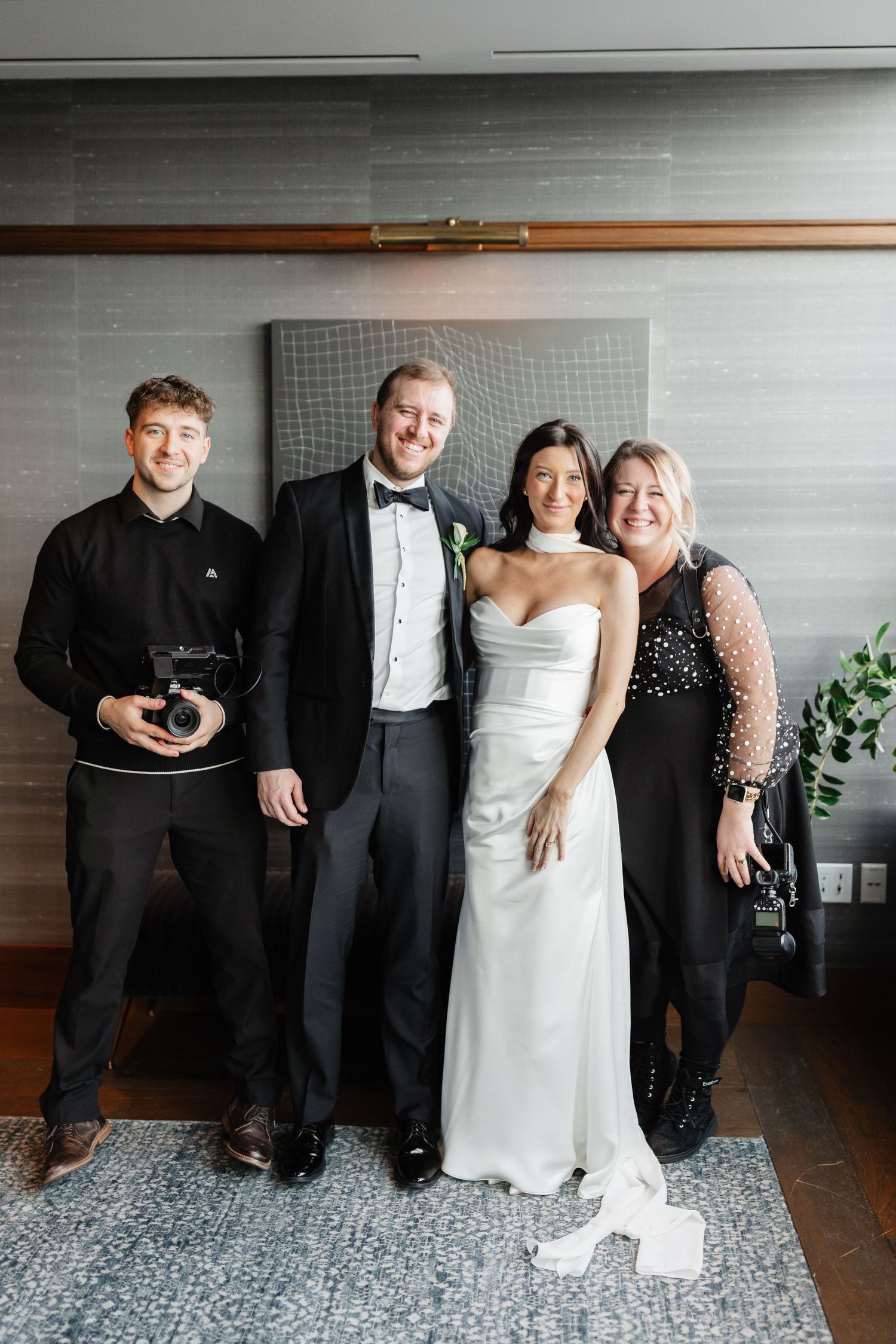 Wedding party of four posing indoors, bride in white gown, groom in tuxedo, guests in formal black attire.
