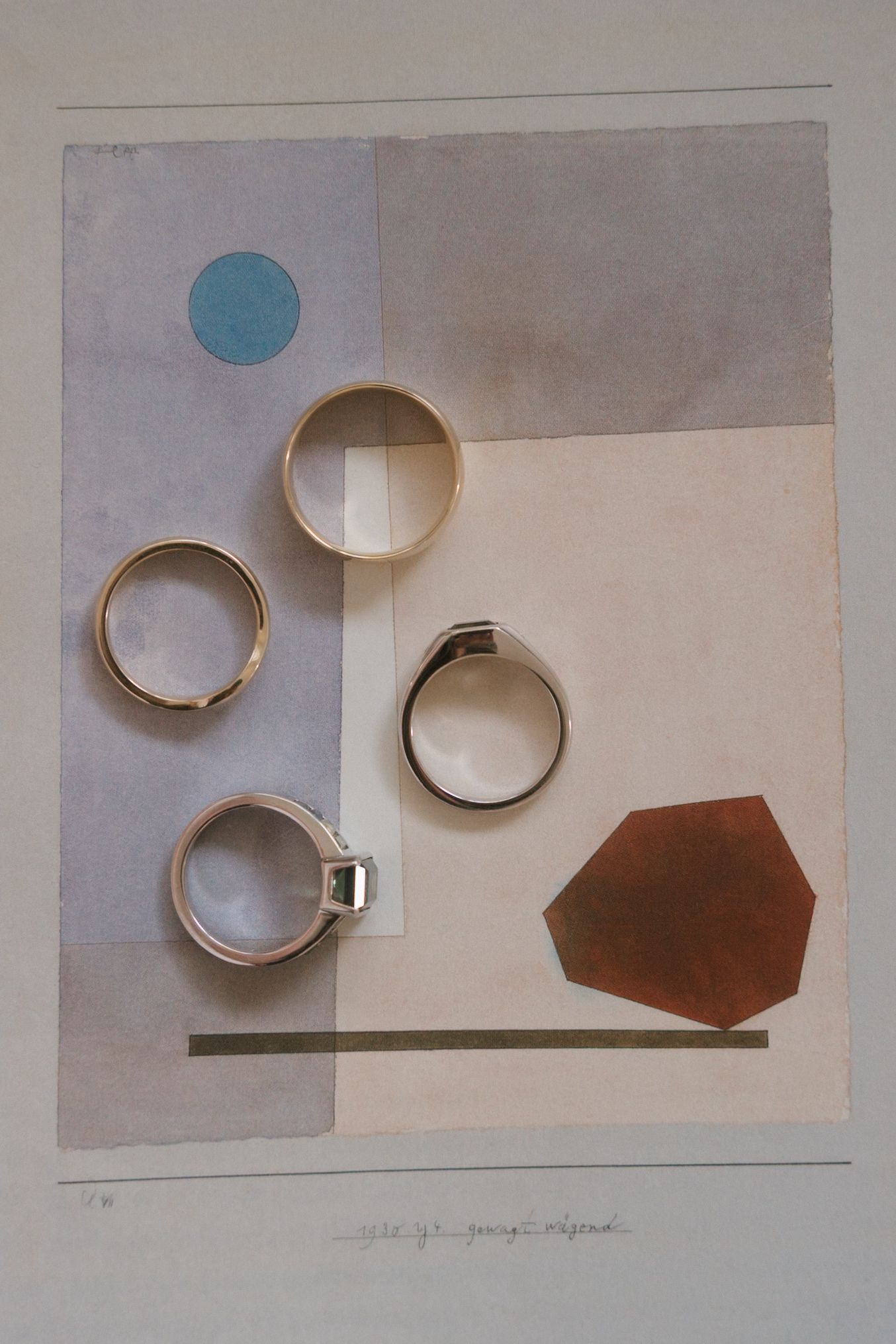 Four gold and silver rings arranged on an abstract geometric collage artwork with blue circle and brown hexagon shapes.