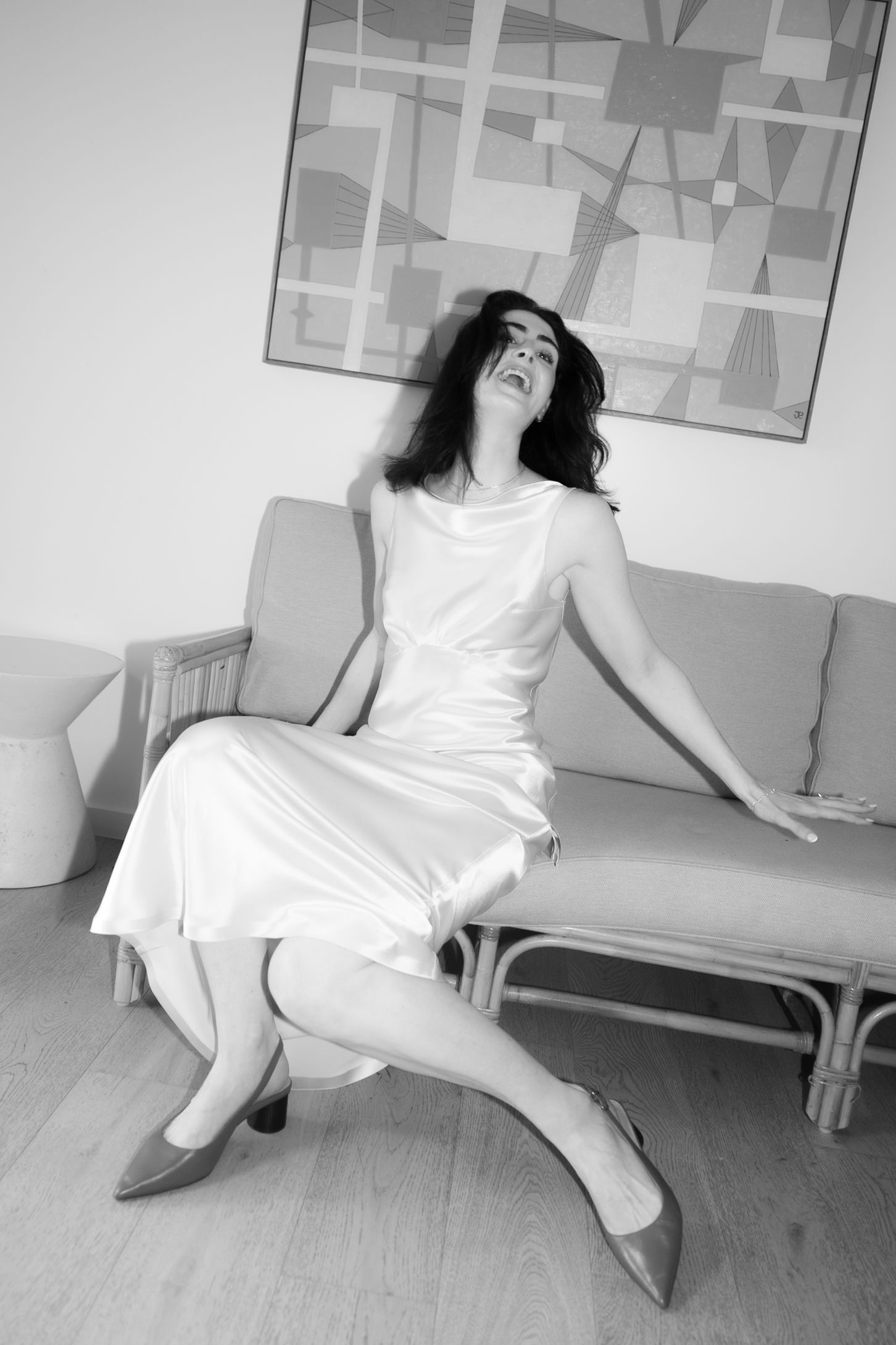 Black and white photo of a woman in a white satin dress sitting on a rattan sofa, head tilted back.