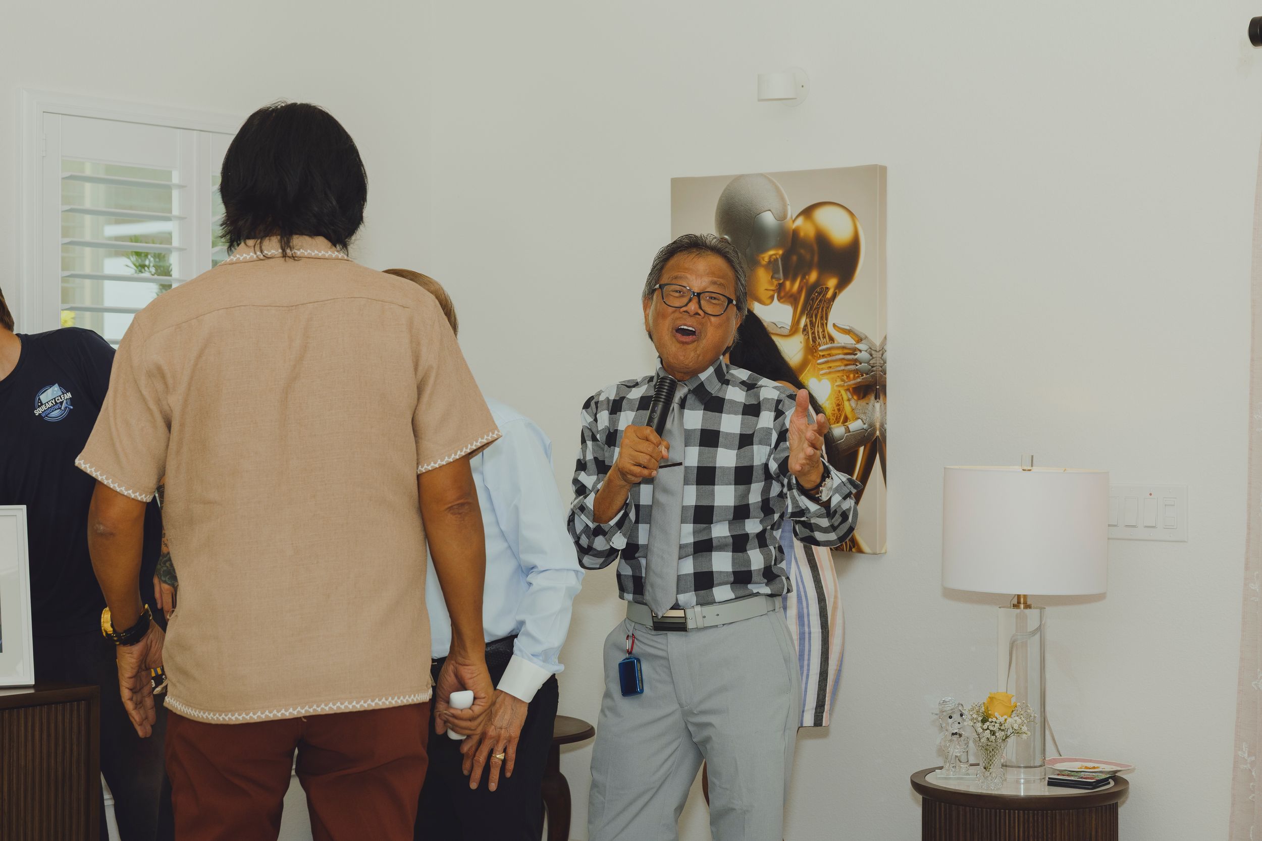 Man in plaid shirt gestures while speaking to guests at an indoor art gallery event.