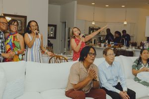 Group of friends and family gathered in a modern home, some standing and singing while others sit on a white sofa.