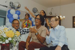 Elderly friends laugh and celebrate at a festive party with blue balloons and flowers in a cozy living room.