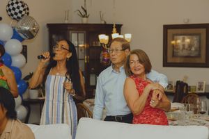 Three people at a festive party, woman holding microphone in striped dress, couple dancing in background with balloon decorations.