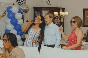 Three adults sing karaoke at a birthday party with blue balloon arch and disco ball decorations.