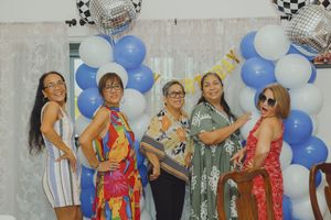 Five women posing together at a celebration with blue and white balloon arch backdrop.
