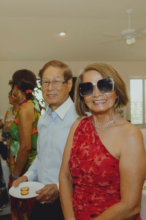 Couple at indoor party, woman in red dress with sunglasses, man in blue shirt holding plate of cookies.