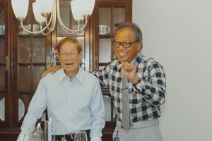 Two elderly Asian men smiling and posing together indoors near a wooden cabinet with glassware.