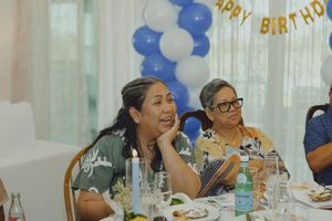 Two women sit at a decorated birthday party table with blue and white balloons and a Happy Birthday banner.