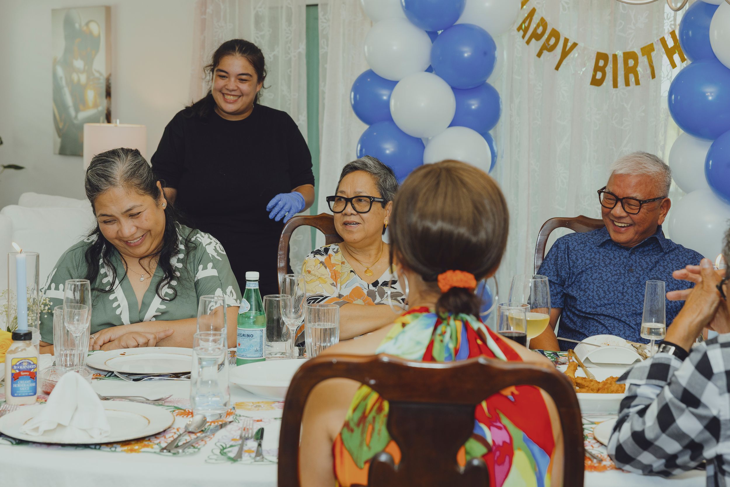 Elderly family and friends gather around a decorated table at a birthday party with blue and white balloon decorations.