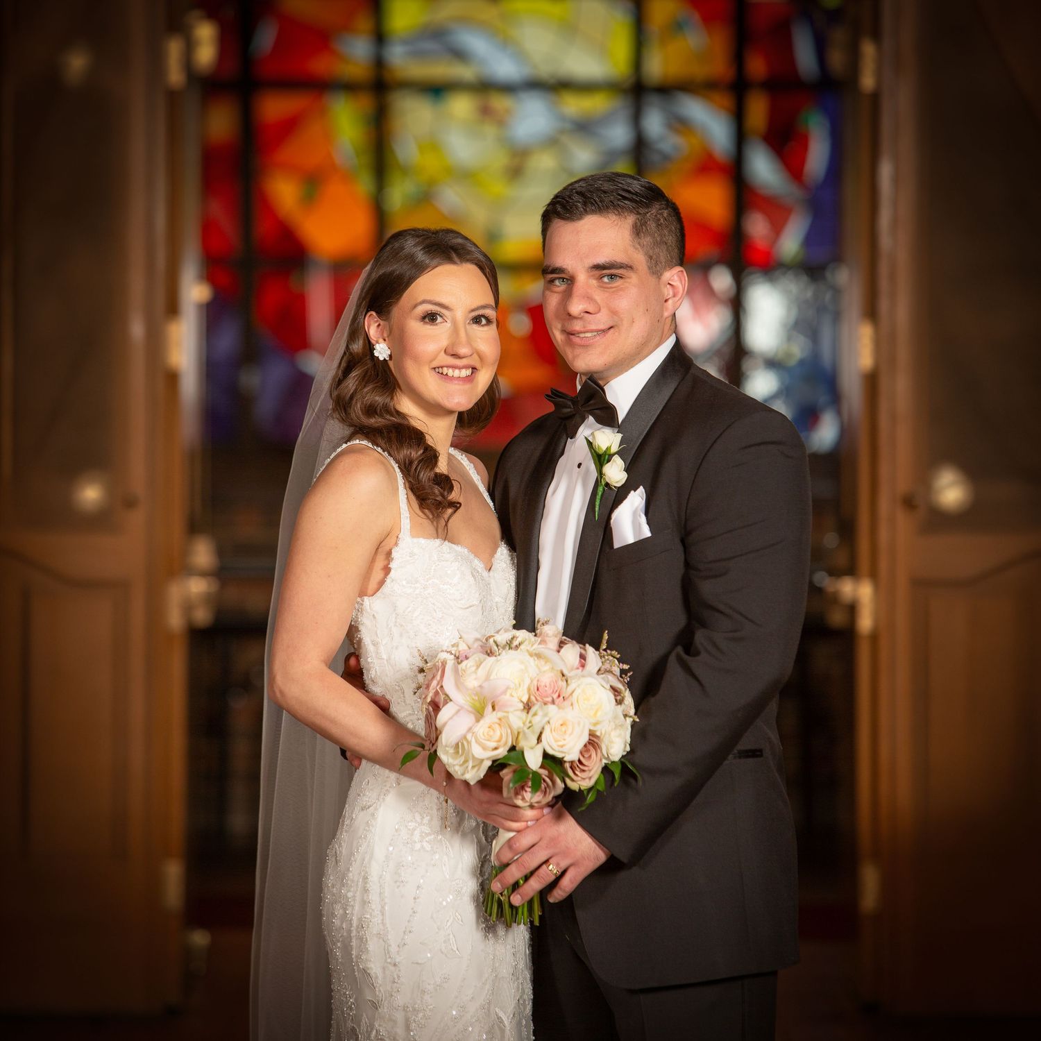 Bride and groom pose elegantly before a vibrant stained glass window, she holds a blush bouquet.