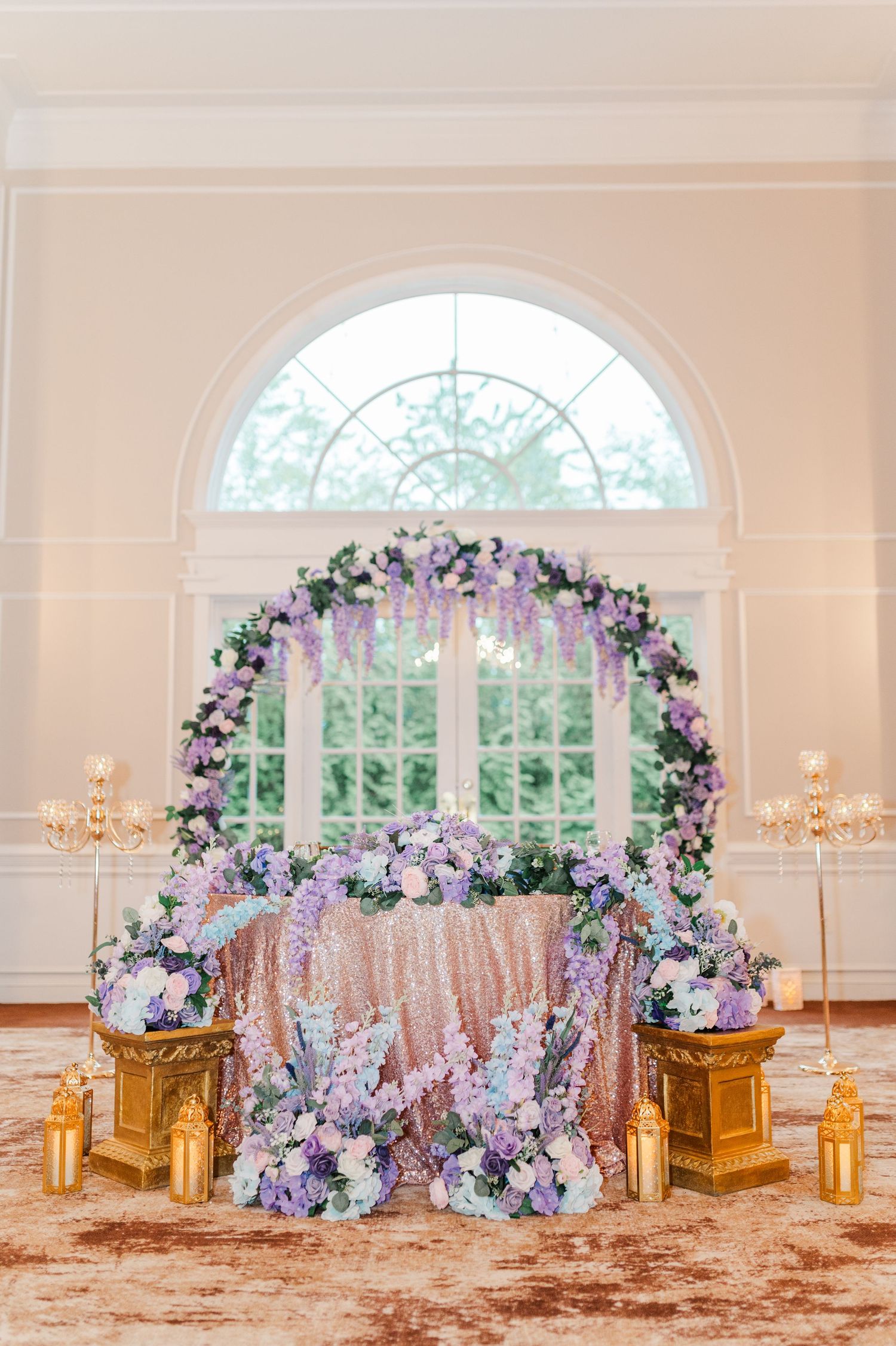 Elegant wedding sweetheart table with lavender floral arch, sequin tablecloth, and gold candle lanterns in a bright ballroom.