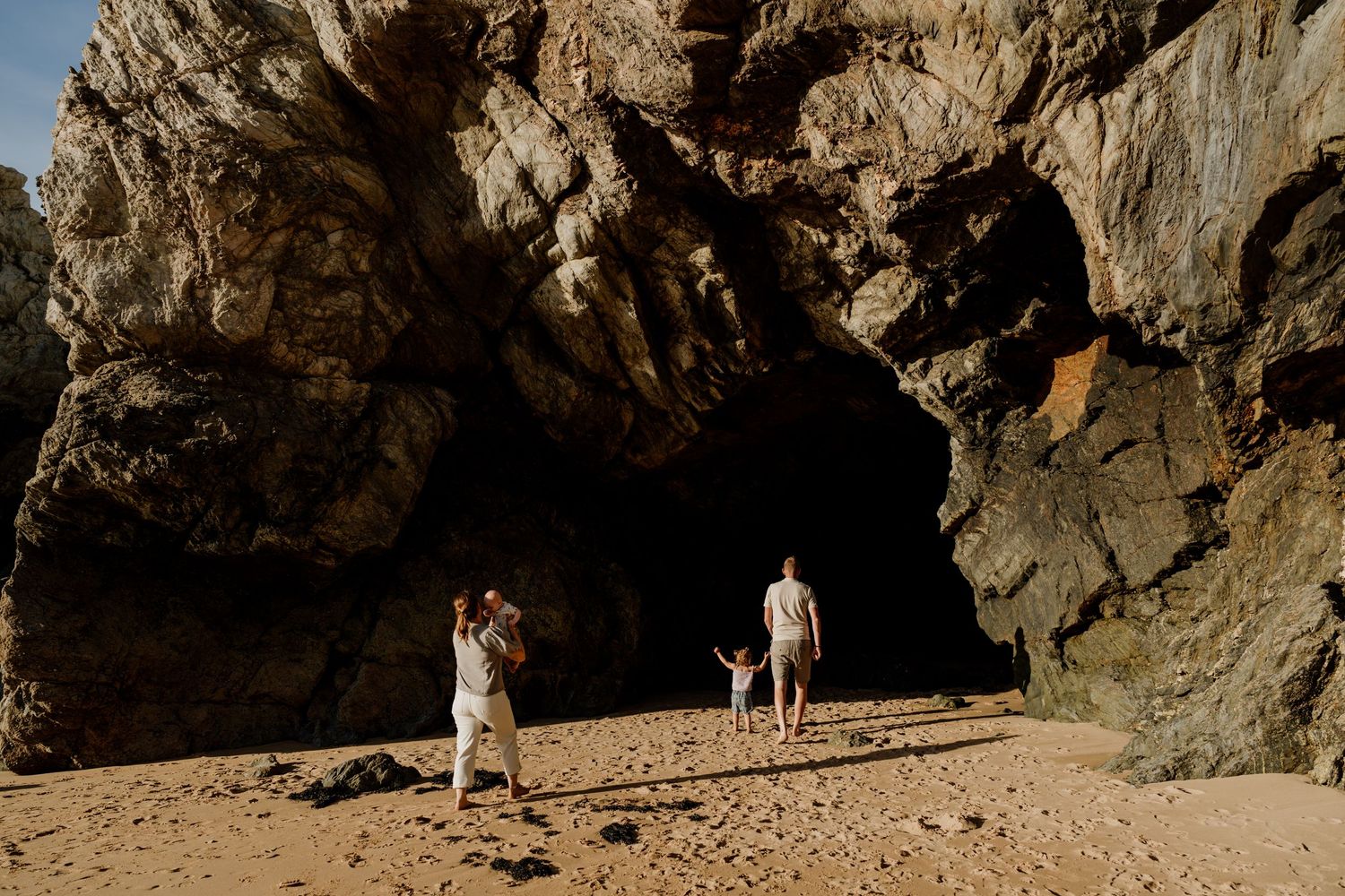 Family of three playing on sandy beach beside dramatic rocky cave formations in golden sunlight.