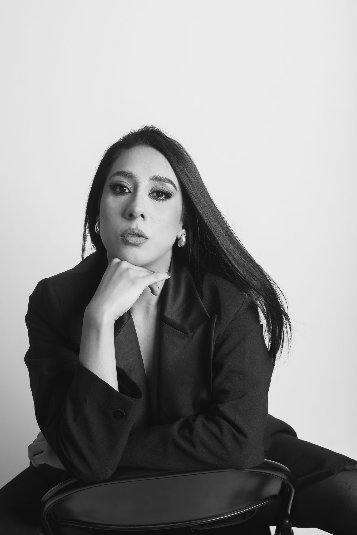 Black and white portrait of a woman in a dark blazer sitting on a chair with hand under chin, elegant pose.