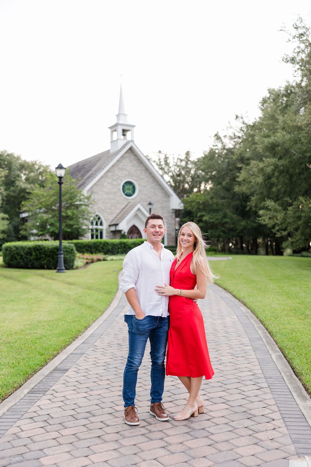 Couple in red dress and white shirt posing on brick path before a stone chapel surrounded by lush greenery.
