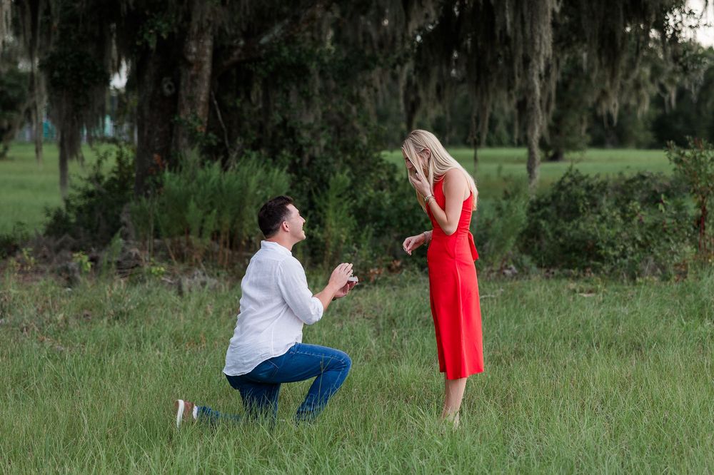 Man kneeling on one knee proposing to woman in red dress outdoors in lush green field under large trees.