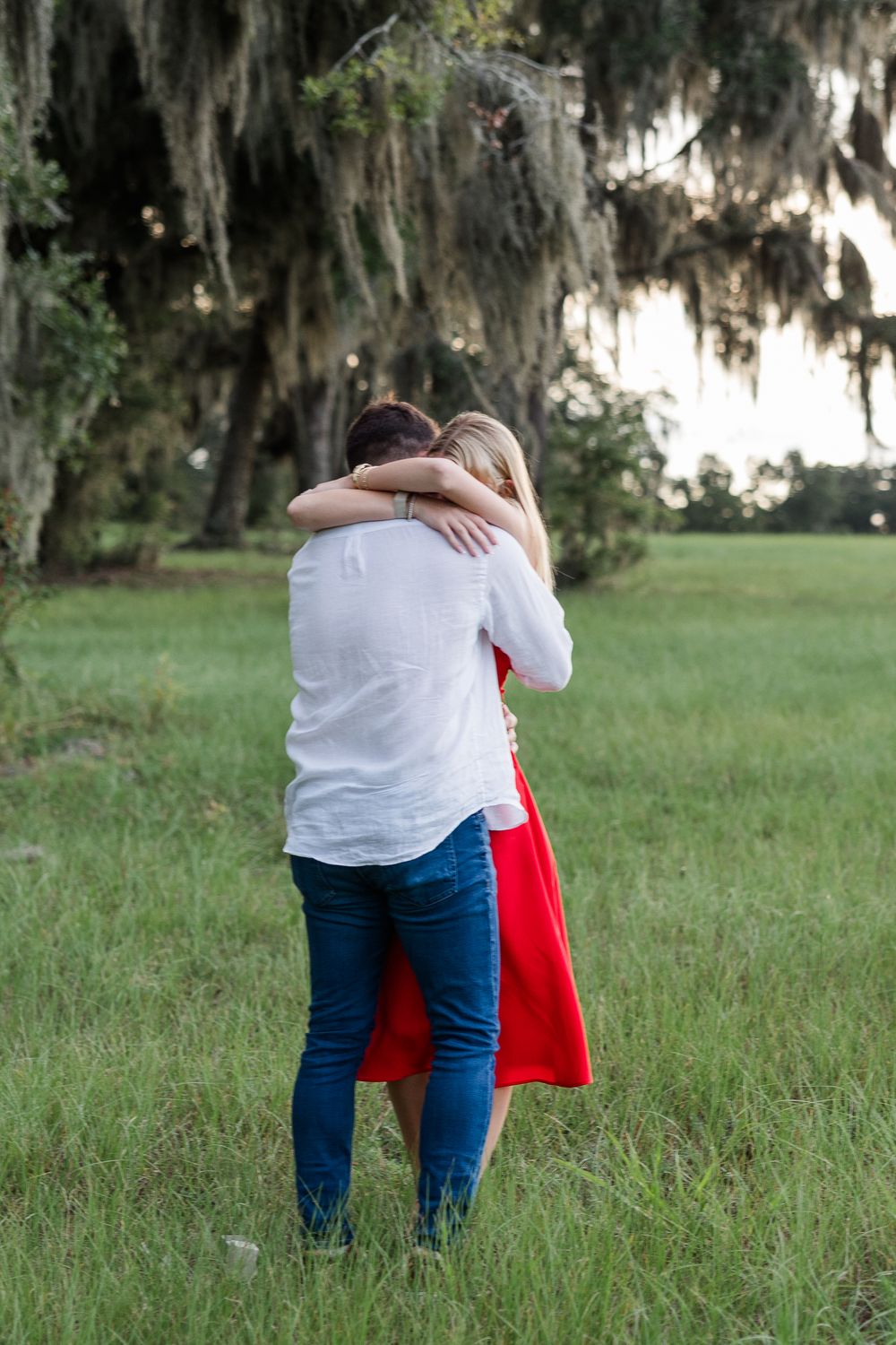 Couple embracing in a lush green field under moss-draped trees, woman wearing a red dress and man in white shirt.