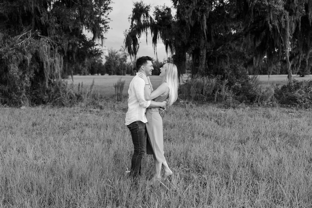 Black and white photo of a couple embracing in a grassy field with weeping trees in the background.