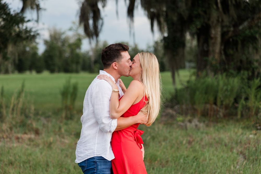 Couple sharing a romantic kiss in a lush green field with Spanish moss-draped trees, woman in red dress.