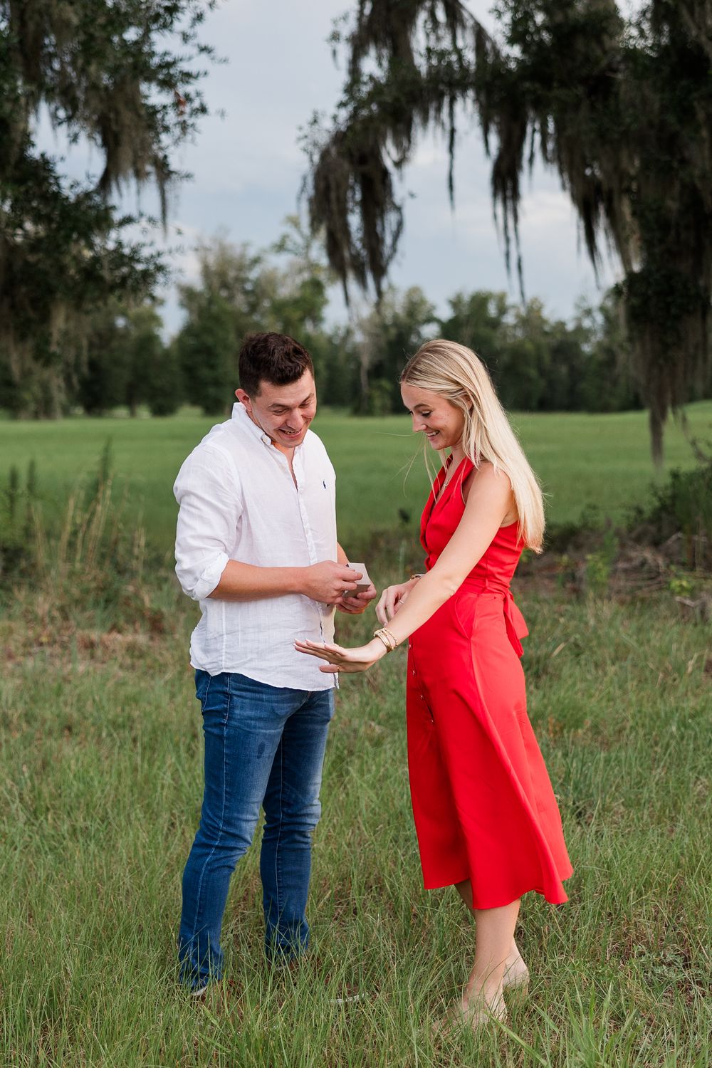 A couple shares a romantic moment in a green field, man in white shirt and jeans, woman in red dress, possibly during a proposal.