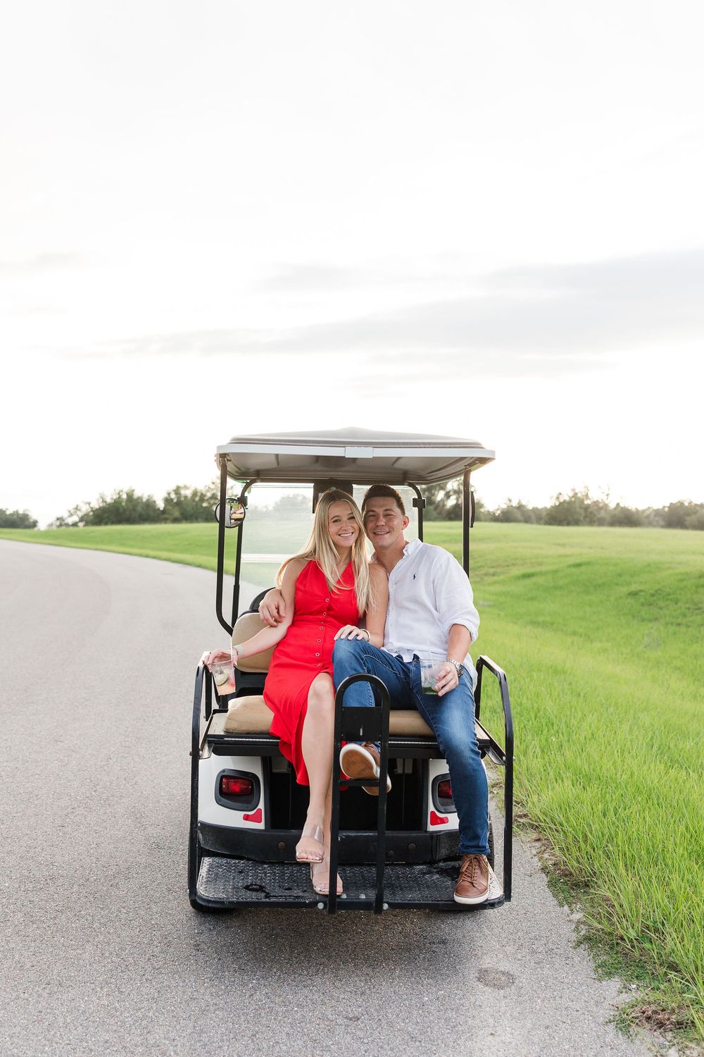 Couple sitting together on the back of a golf cart on a path surrounded by green grass.