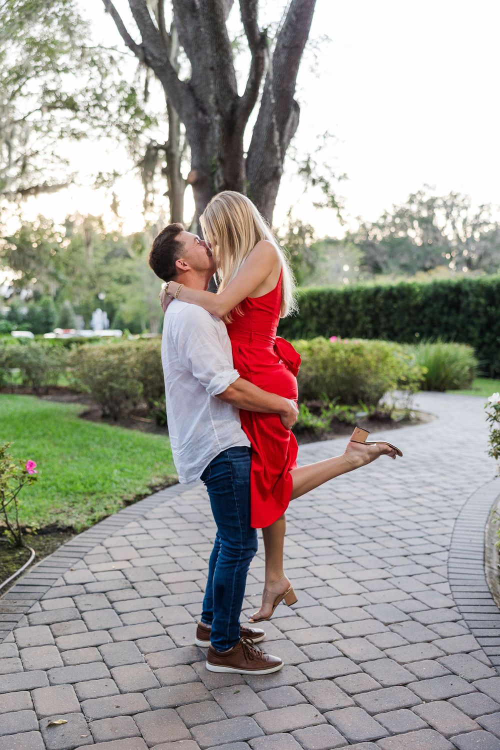 A couple shares a romantic kiss as the man lifts the woman in a red dress on a brick garden path.