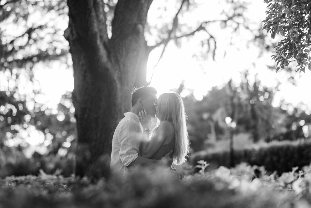 Black and white photo of a couple sharing a romantic kiss under a large tree in a lush garden setting.