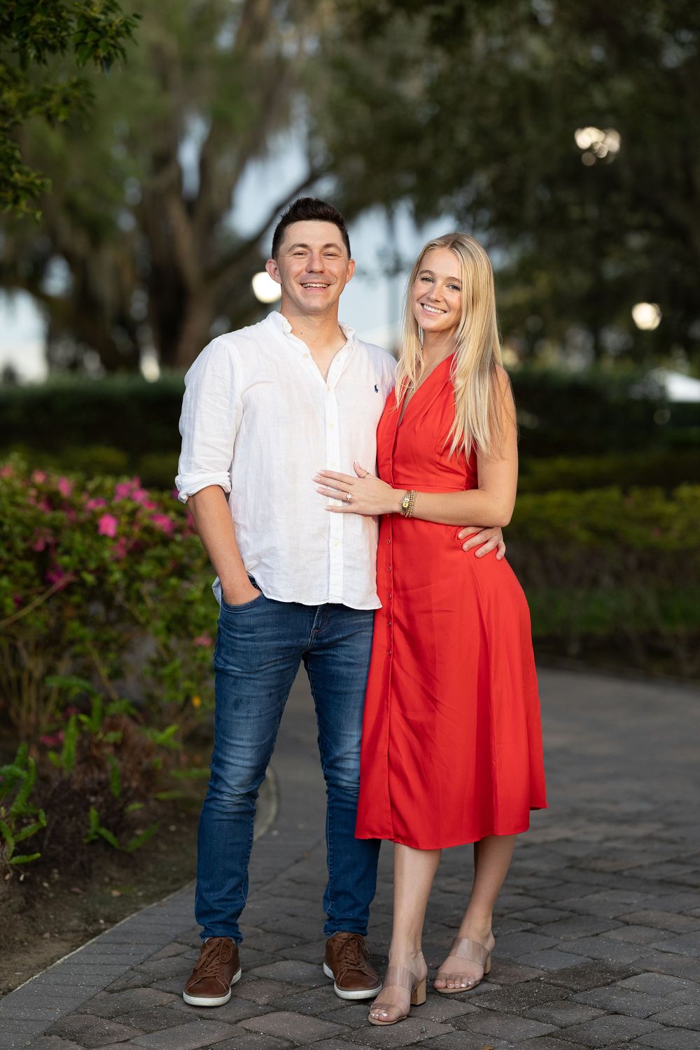 Smiling couple posing outdoors, man in white shirt and jeans, woman in red dress, lush green garden background.