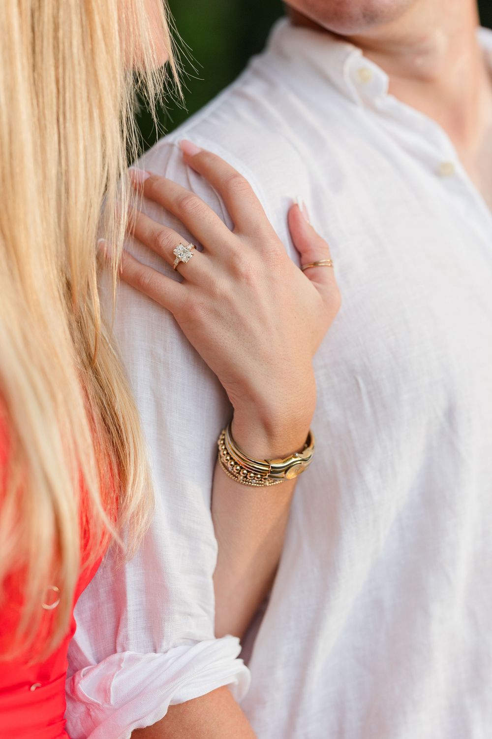 Close-up of a woman's hand with a diamond engagement ring and gold bracelets resting on a man's white linen shirt.