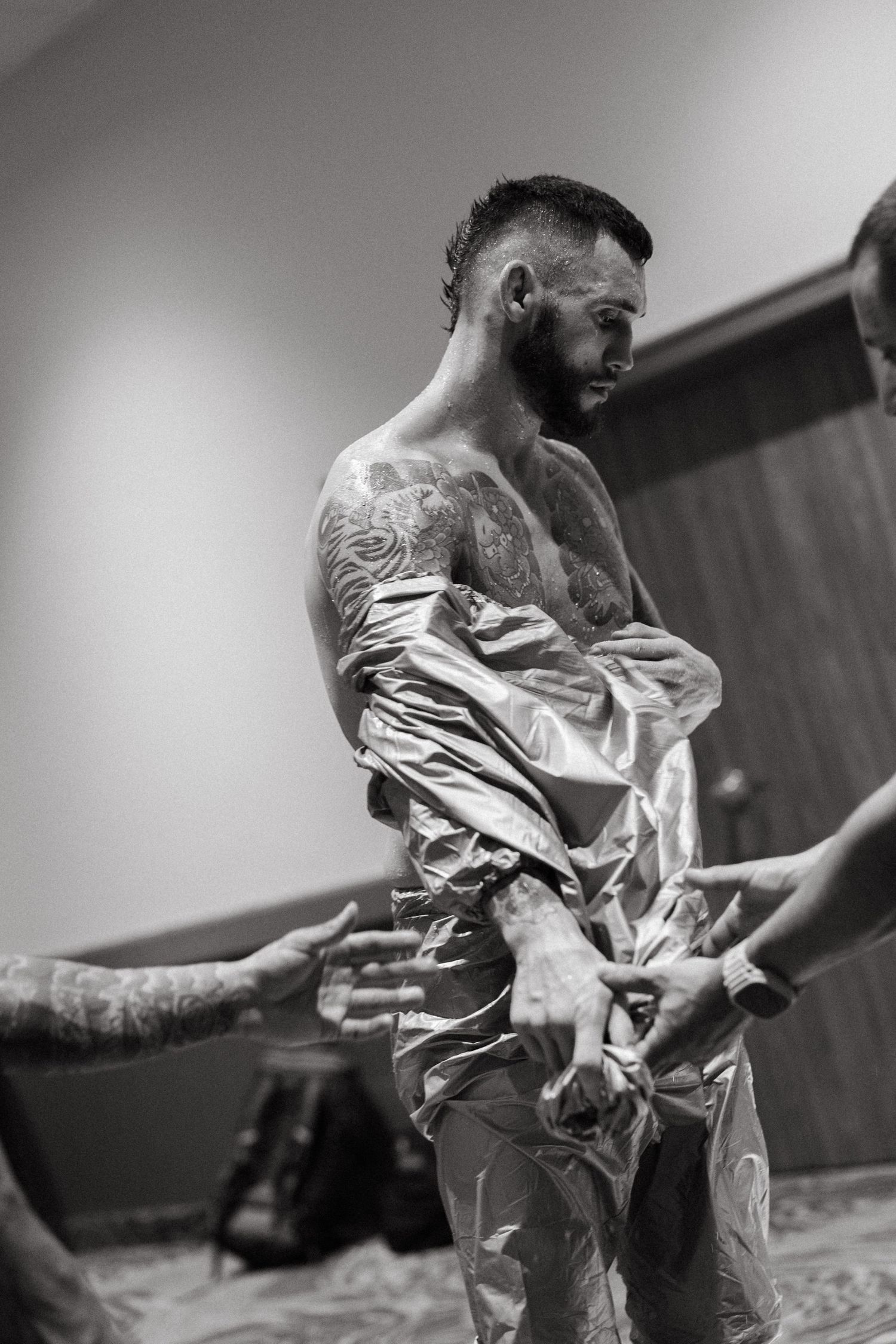 Black and white photo of tattooed shirtless man being helped into shiny pants by others in a room.
